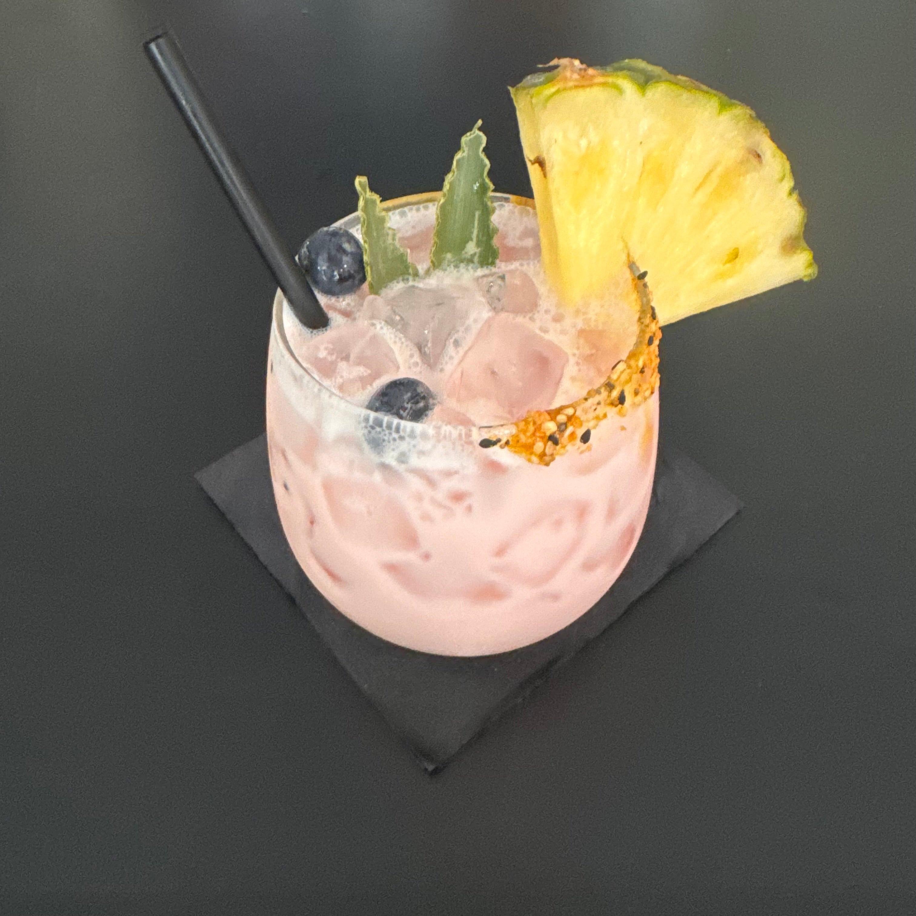 Pink cocktail with pineapple slice and black straw on a dark surface with sloan seasonings citrus sriracha on the rim.
